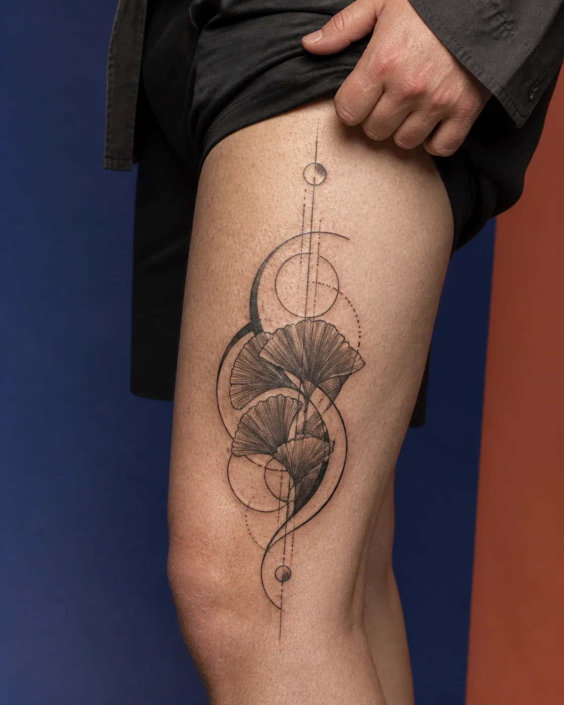 Geometric tattoo of overlapping ginkgo fans carried by a curved stem executed as a healed cover-up on the outer thigh