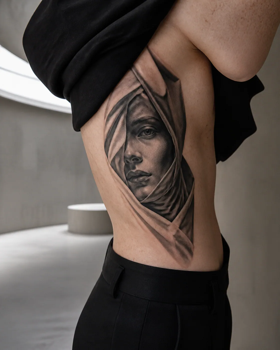 Black and Gray tattoo of an androgynous face emerging from folded fabric on the side ribs
