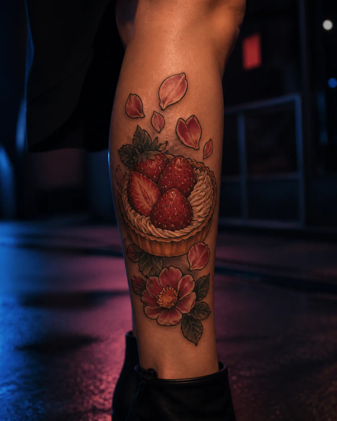 Neo-Traditional tattoo of a strawberry pastry surrounded by scattered petals executed as a healed cover-up on the calf