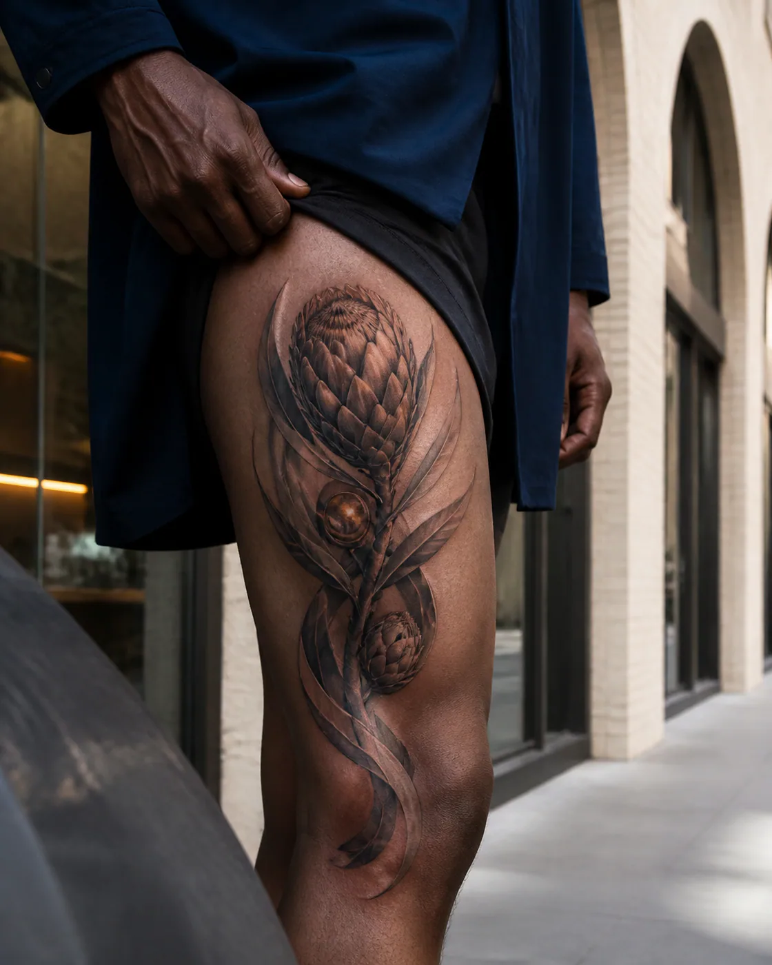 Surrealism tattoo of a sculptural king protea opening between silver leaves with a three-part family motif on the outer thigh