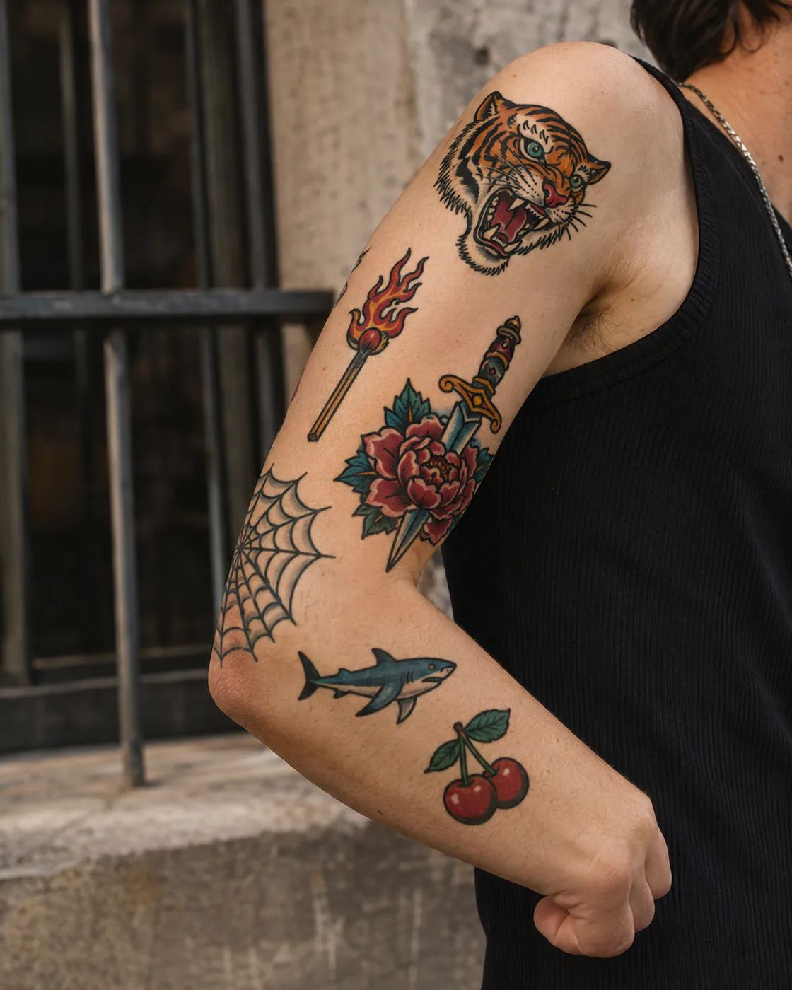 Bent-arm American traditional patchwork sleeve tattoo with a tiger, peony, dagger, web, shark, match, and cherries on light skin