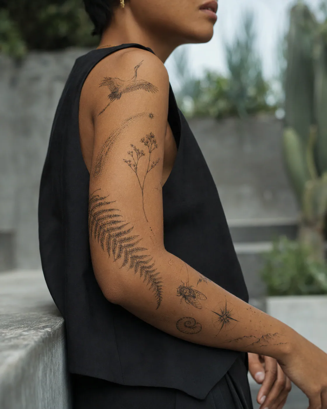 Bent-arm fine-line patchwork sleeve tattoo with a flying crane, arcing fern, comet, wildflowers, cicada, shell, and mountain line on brown skin