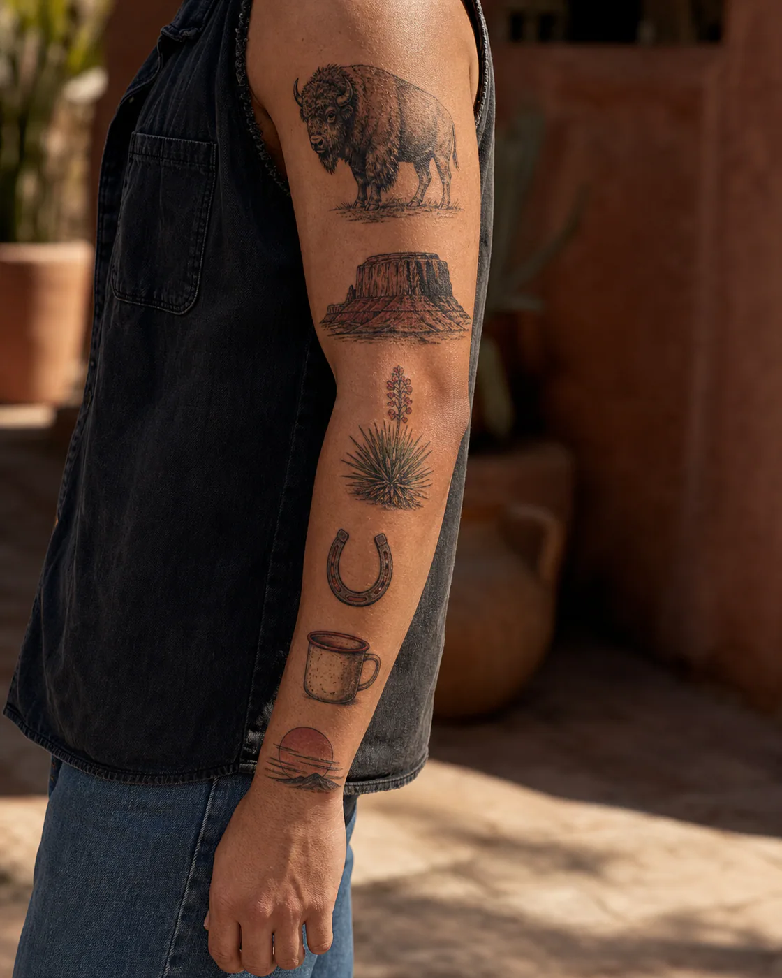 Western illustrative patchwork sleeve tattoo with a bison, desert mesa, yucca bloom, horseshoe, camp mug, and setting sun on olive skin