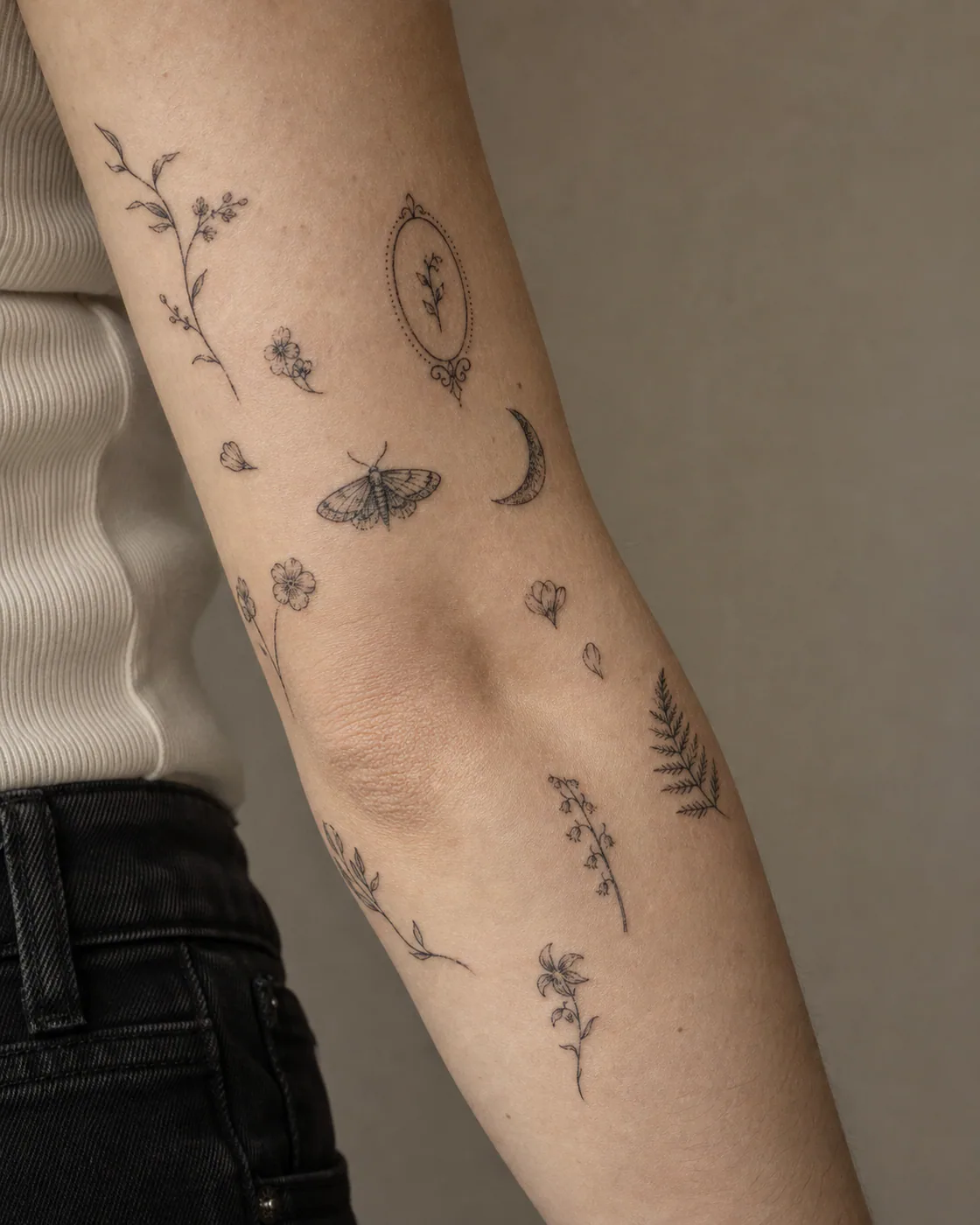 Fine-line patchwork sleeve tattoo on light skin with small blossoms, curved stems, fern tips, petals, and intentional gaps flowing around existing elbow pieces